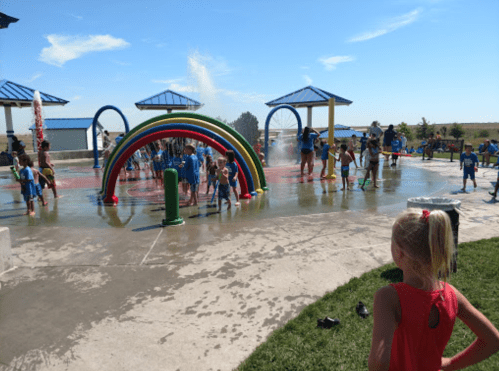 Children play in a colorful splash pad with water features on a sunny day, surrounded by grassy areas and blue skies.