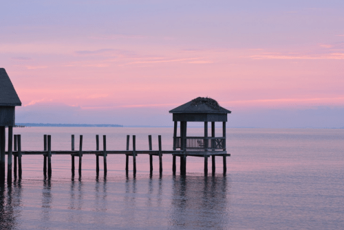 A serene waterfront scene at sunset, featuring a wooden pier and a gazebo with a nest, reflecting on calm waters.