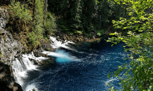 A serene blue river with small waterfalls, surrounded by lush green trees and rocky terrain.