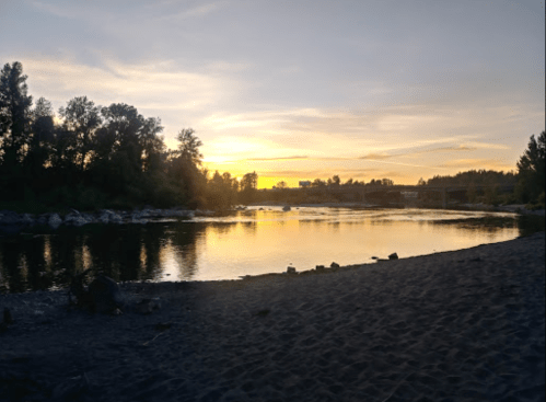 A serene river at sunset, with trees lining the shore and a sandy beach in the foreground.