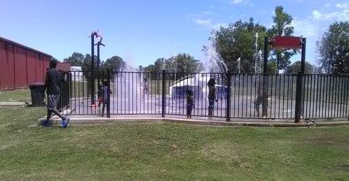 Children play in a splash pad, running through water jets on a sunny day, surrounded by a black fence and grassy area.