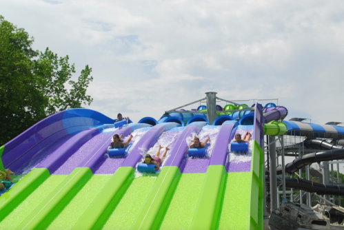 Four people slide down colorful water slides, splashing into a pool below, surrounded by greenery and cloudy skies.