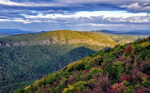 A panoramic view of lush green mountains under a cloudy sky, with hints of autumn colors in the foreground.