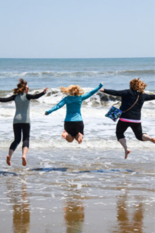 Three women jump joyfully at the beach, splashing in the waves with the ocean in the background.