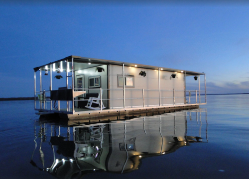 A modern houseboat illuminated at dusk, floating on calm water with reflections visible.