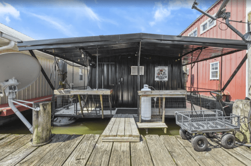 A black houseboat with a covered deck, wooden walkway, and nearby red buildings on a calm waterway.