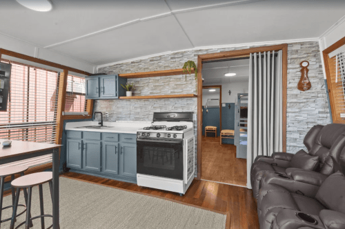 A modern kitchen with blue cabinets, a gas stove, and a cozy seating area, featuring wooden floors and stone accents.
