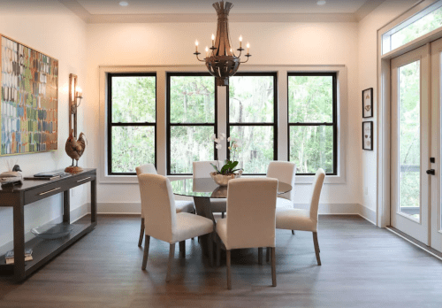 A modern dining area with a round table, white chairs, a chandelier, and large windows overlooking greenery.