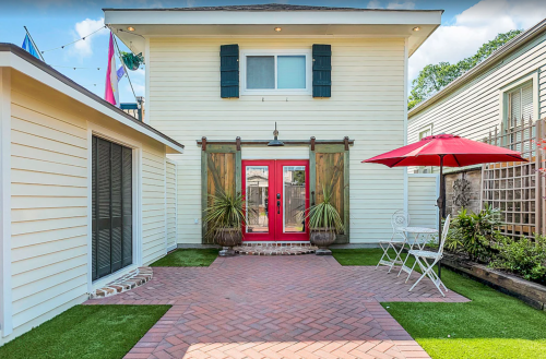 A charming outdoor space featuring a brick patio, red doors, potted plants, and a red umbrella over a small table.