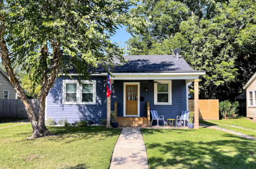 A blue house with a front porch, two chairs, and a pathway, surrounded by green grass and trees on a sunny day.