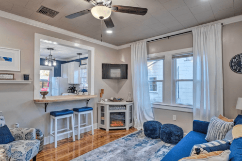 Cozy living room with a blue sofa, decorative pillows, a small bar area, and a fireplace, featuring light walls and wooden floors.