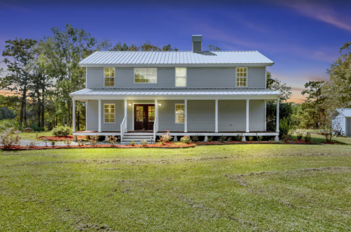 A two-story gray house with a porch, surrounded by greenery and flower beds, under a twilight sky.