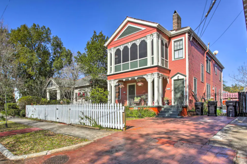 A charming red Victorian house with a white picket fence and lush greenery, set on a sunny street.
