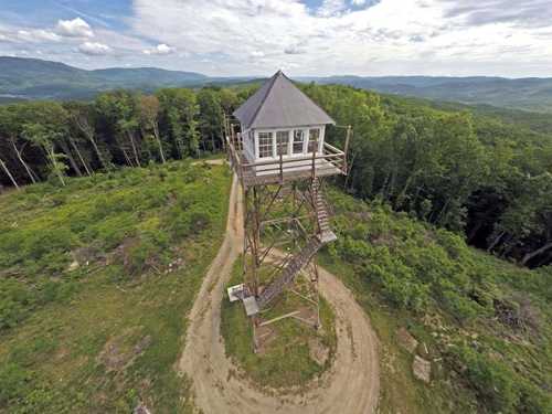 A tall wooden observation tower surrounded by lush green trees and rolling hills under a cloudy sky.