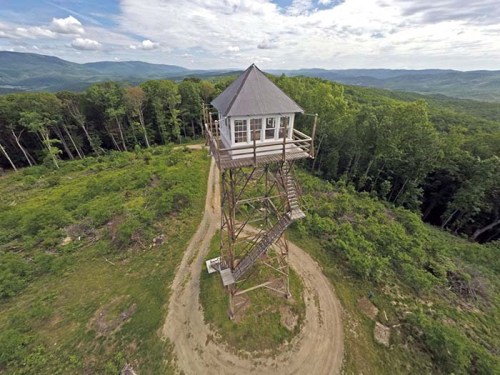 A tall wooden observation tower surrounded by lush green trees and rolling hills under a cloudy sky.