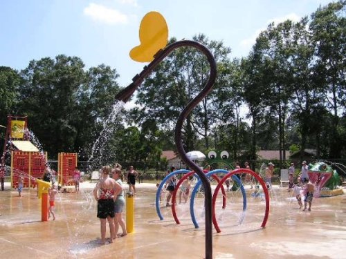 A vibrant splash pad with colorful water features and children playing in the sun, surrounded by trees.