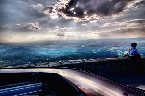 A person sits on a stone wall, gazing at a scenic view of mountains and valleys under a dramatic sky.