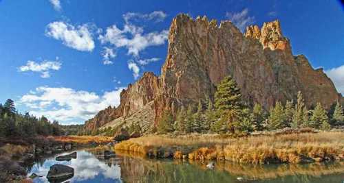 A scenic landscape featuring towering rock formations beside a calm river, surrounded by trees and blue skies.