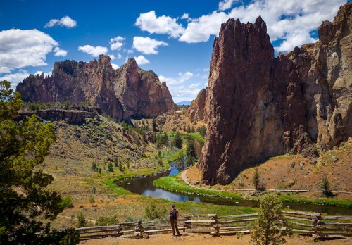 A person stands near a river surrounded by towering rock formations and lush greenery under a blue sky with clouds.