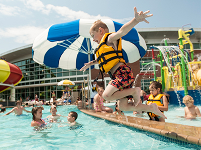 A child in a life jacket jumps into a pool, surrounded by other kids and colorful water attractions on a sunny day.