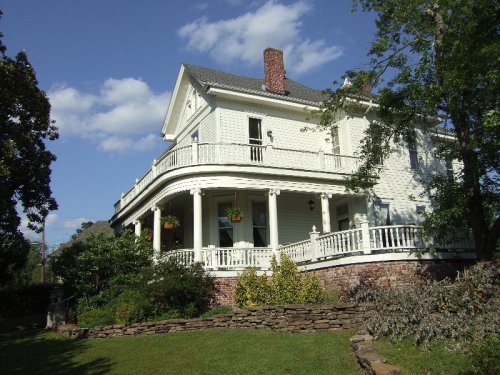 A large white Victorian-style house with a wraparound porch, surrounded by greenery and under a blue sky.