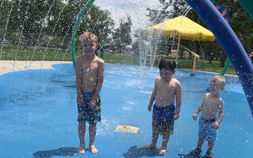 Three children play in a splash pad, enjoying water sprays on a sunny day.