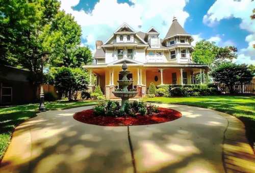 A charming Victorian-style house with a fountain in the foreground, surrounded by lush greenery and blue skies.