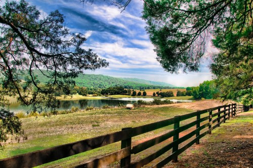 A serene landscape featuring a lake, green hills, and a wooden fence under a blue sky with scattered clouds.