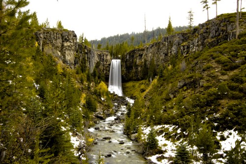 A serene waterfall cascades down rocky cliffs, surrounded by lush greenery and a winding river below.