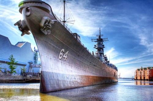 A large battleship with the number 64 on its bow, docked near modern buildings under a bright blue sky.