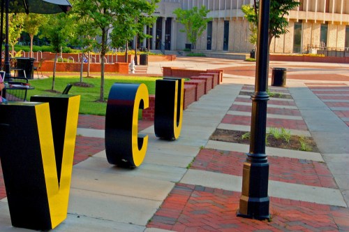 Large yellow letters spelling "VCU" in a landscaped outdoor area with trees and benches.