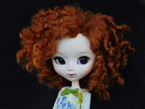 A close-up of a doll with curly red hair and large eyes, wearing a floral dress with a green bow.