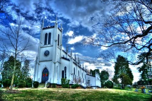 A white church with tall spires, surrounded by trees and a cloudy sky, set in a peaceful landscape.