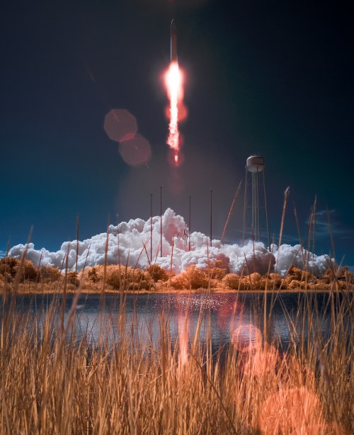 A rocket launches into the sky, leaving a trail of smoke over a calm lake surrounded by tall grass.