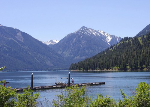A serene lake surrounded by mountains, with a dock and a person near a boat under a clear blue sky.