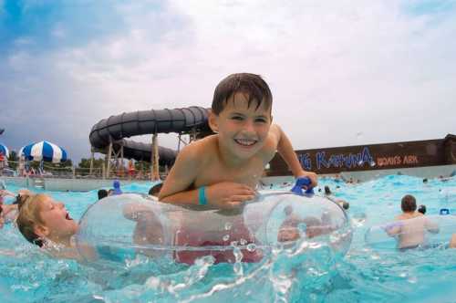 A smiling boy floats on a tube in a water park, surrounded by other children and water attractions.