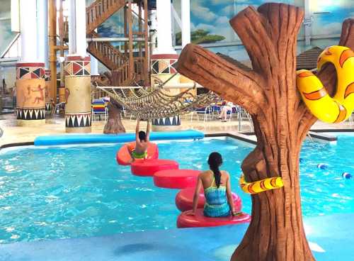 Two children play on floating steps in a pool, with a tree sculpture and a colorful snake decoration nearby.