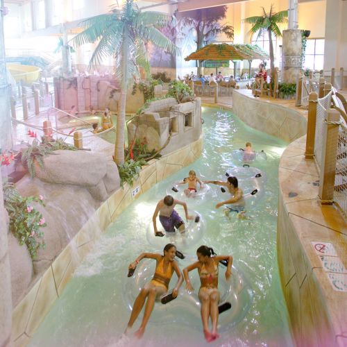 A water park scene with people floating on inner tubes in a lazy river surrounded by tropical plants and rocks.