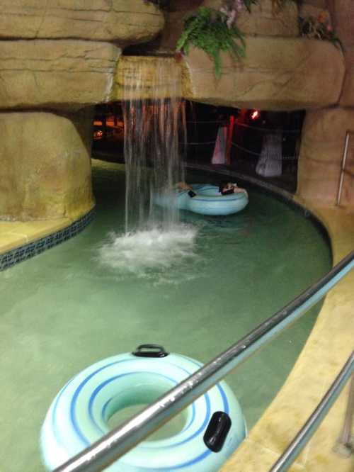 Two blue inner tubes float in a winding pool beneath a waterfall, surrounded by rocky decor and greenery.