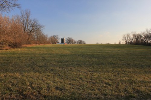 A wide, open field with sparse trees and a distant farm structure under a clear sky.