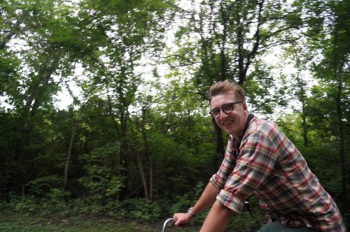 A person with glasses smiles while riding a bicycle through a lush, green forested area.