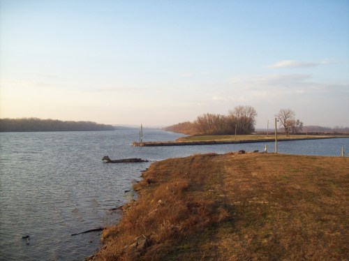 A serene river scene with grassy banks, trees in the distance, and a clear sky reflecting on the water.