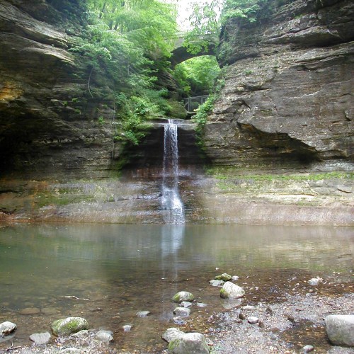 A serene waterfall cascades into a calm pool, surrounded by rocky cliffs and lush greenery.