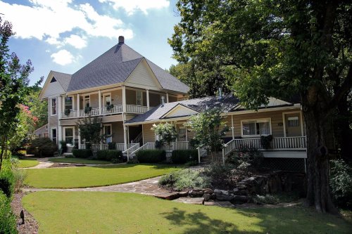 A large, two-story house with a wraparound porch, surrounded by greenery and a well-maintained lawn under a blue sky.