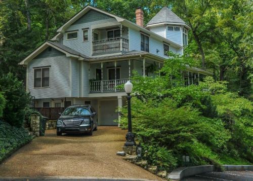 A large, two-story house surrounded by trees, featuring a driveway and a parked car in front.