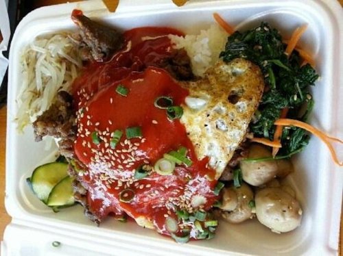 A takeout container filled with rice, meat, vegetables, an egg, and a red sauce, garnished with green onions and sesame seeds.