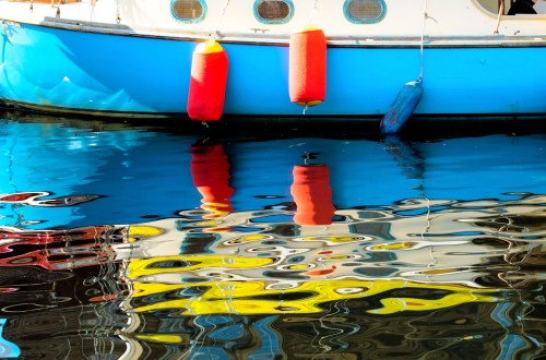 A colorful boat with orange buoys reflects in calm water, creating vibrant patterns and ripples.