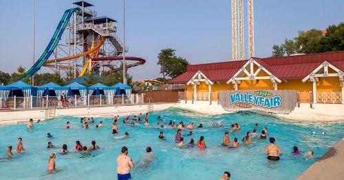 A busy water park scene with people swimming in a pool, colorful slides in the background, and sunny skies.