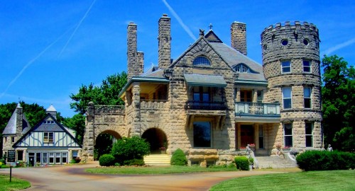 A large, historic stone mansion with towers and a turret, surrounded by green lawns and blue skies.