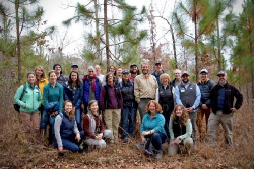 A diverse group of 30 people poses together in a forested area, surrounded by pine trees and autumn foliage.